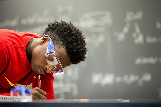 图为英国波胆网站的一名学生在实验室里穿着红色衬衫，戴着护目镜, writing with a pencil. 